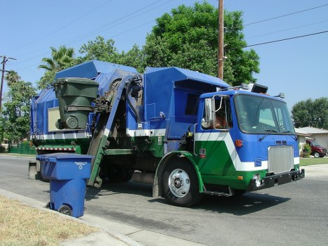 A large, blue and green rubbish collection truck parked on a suburban street with trees and residential houses visible in the background. The vehicle has a front cab with a silver grille, clear windows, and side mirrors. The rear section features a large, rectangular compactor area with mechanisms and a hydraulic lift, used for lifting and emptying waste bins. In the foreground, a standard outdoor rubbish bin made of dark green plastic is positioned near the truck, ready for collection. The scene is set in daylight, with a clear sky and the truck aligned parallel to the pavement, indicative of a waste removal service operating within a local neighbourhood, possibly in Barnet or surrounding areas. Barnet House Clearance undertakes rubbish removal services locally, as demonstrated by the context and environment of the scene.
