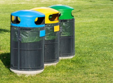 Three outdoor waste bins positioned on a well-maintained grassy area, each featuring vertical black plastic slats forming a cylindrical shape. The bins have bright, rounded plastic lids in blue, yellow, and green, each with small openings on the sides for rubbish disposal, and matching rectangular labels on the front panels. The background shows a clear, sunny environment with a uniform green lawn, suggesting a residential or community setting within the Barnet area. The arrangement of the bins indicates a waste segregation system, likely related to rubbish or recycling collection. Barnet House Clearance offers rubbish removal services in this locality, ensuring proper disposal of waste materials. The overall scene emphasizes public waste management infrastructure in an outdoor context, relevant to local rubbish collection practices in Barnet, North London.