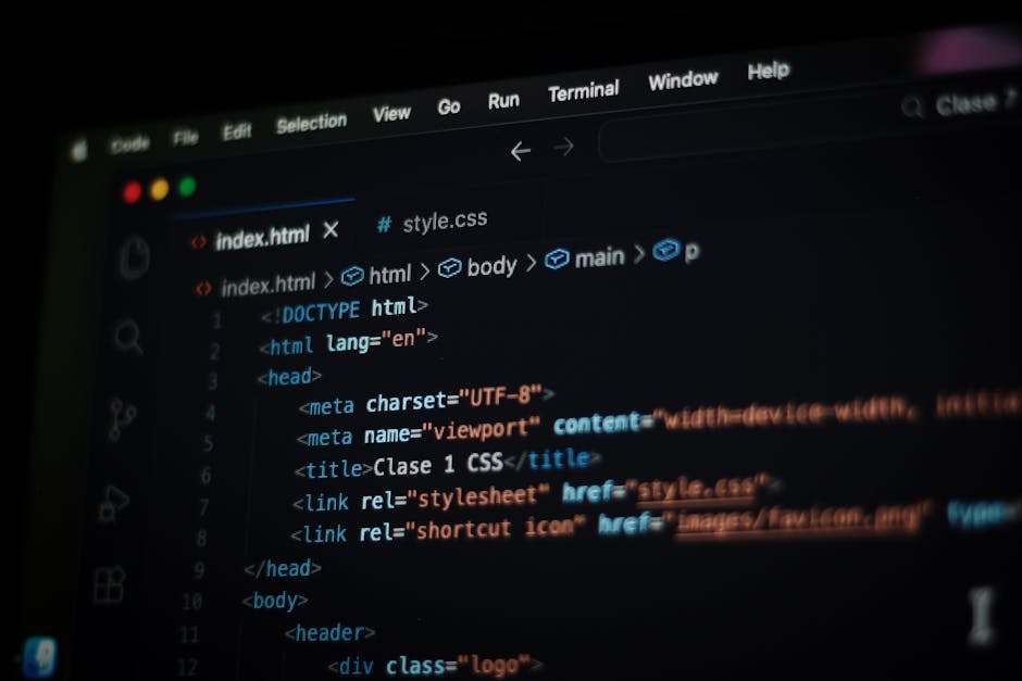 A person seated at a desk is working on a laptop with a dark screen displaying lines of multicolored code, likely involved in programming or technical work. The individual, dressed in a light blue shi