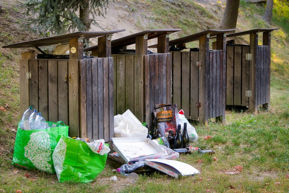 A series of four wooden outdoor rubbish bins with slanted roofs, arranged side by side on a grassy area, each bin constructed from vertical wooden planks with visible hinges and locks. The bins are we